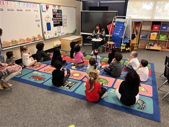 Students are on the carpet listening to their teacher reading a book